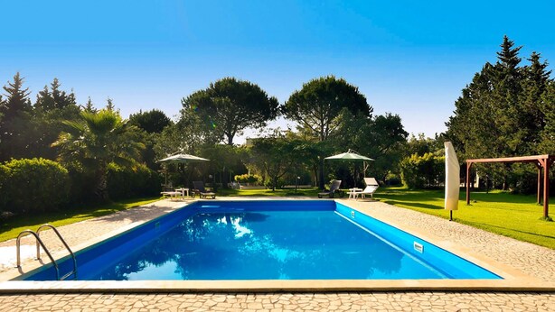 A rectangular swimming pool with a stone deck surrounding it, set in a landscaped garden with trees and umbrellas.