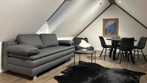 A modern loft bedroom with a sloped ceiling, a grey sofa, a dining table with chairs, and a large window. - 4 Elements Resort Bled Studio Standard (Radovljica)