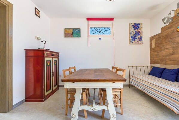 A dining area with a wooden table and chairs, a red cabinet, and a bed with a striped blanket.