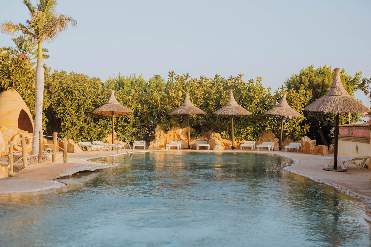A swimming pool with lounge chairs and thatched umbrellas, surrounded by greenery and a stone wall.