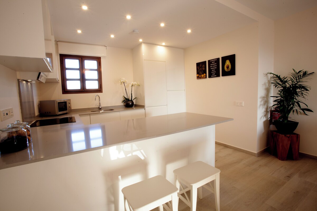 A modern kitchen with a central island, white bar stools, a microwave, and a potted plant.
