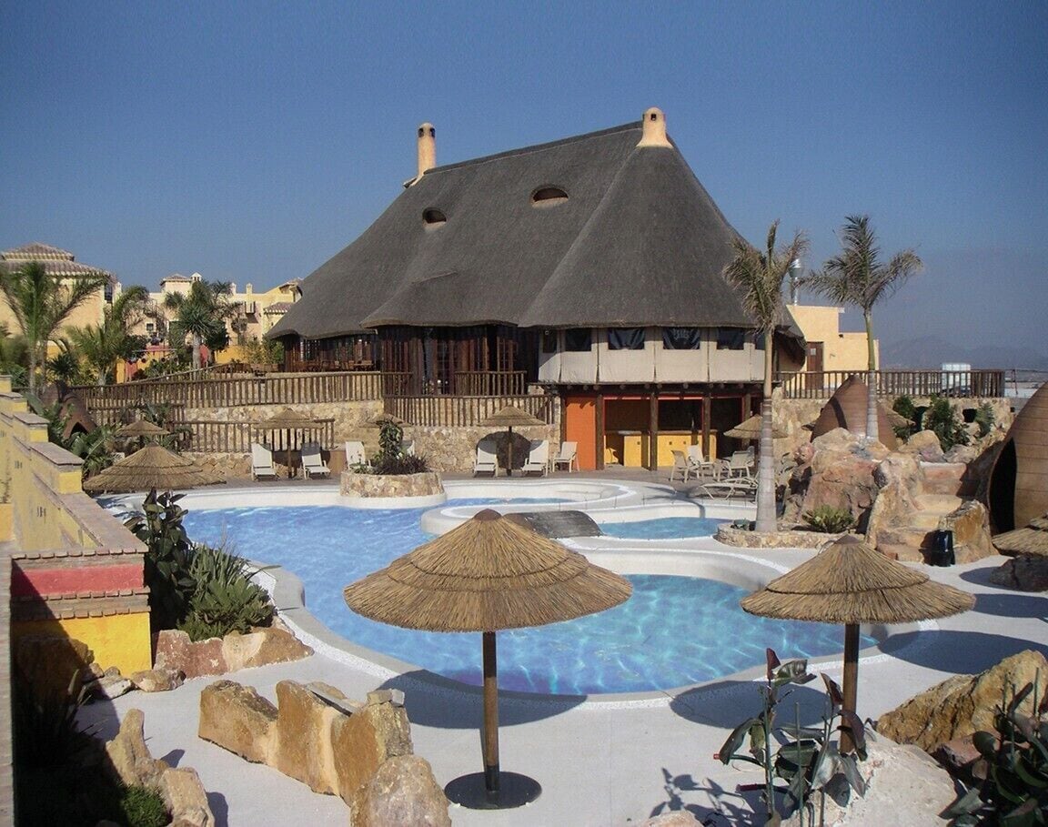 A resort with a large pool, thatched umbrellas, and a building with a dark roof.