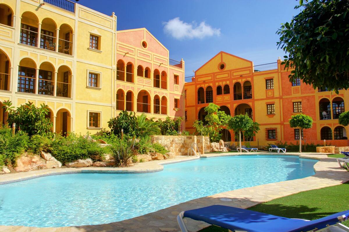 Zona de piscina con una piscina azul cristalina, rodeada por un edificio con varios balcones y un muro de piedra.