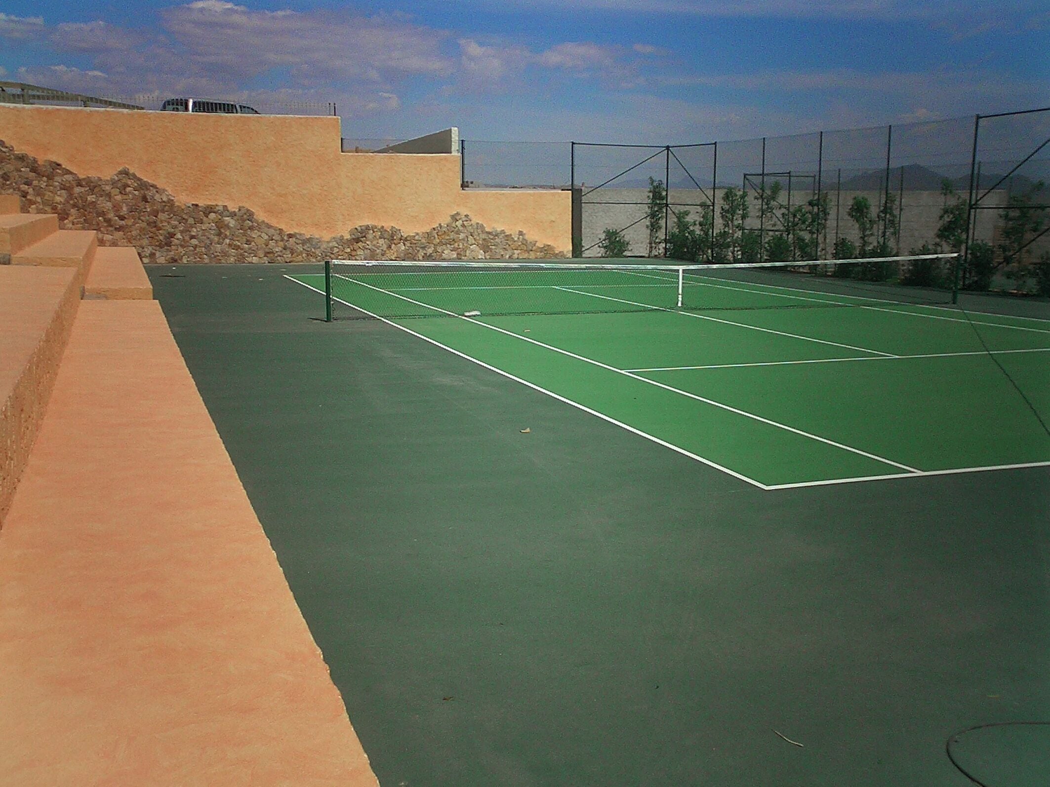 Una cancha de tenis con red, rodeada por un muro bajo y un muro de piedra.