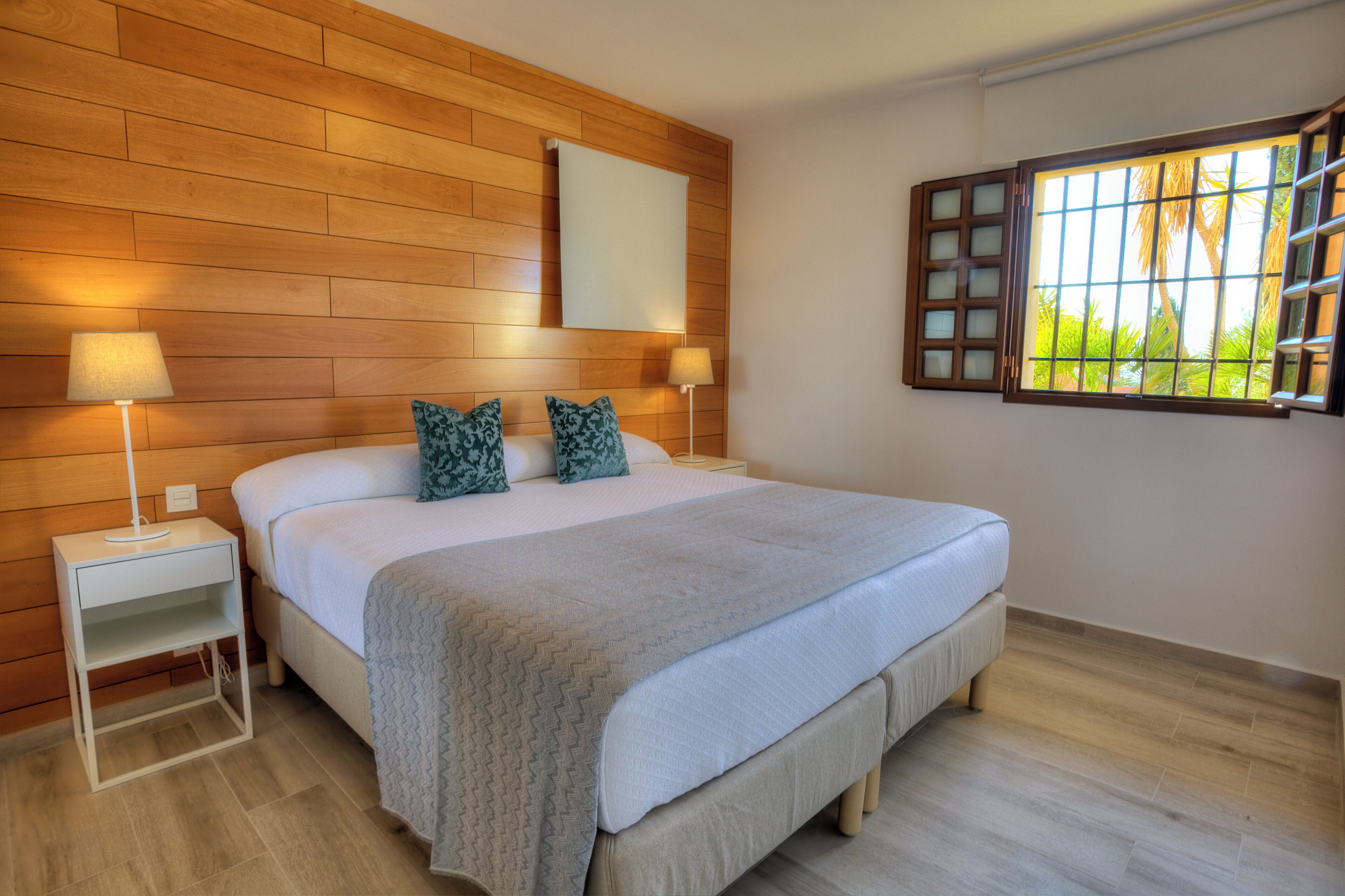Un dormitorio con una cama grande, dos mesitas de noche con lámparas, un cabecero de madera, una ventana con persianas y vistas a zonas verdes.