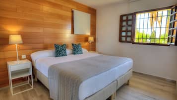 Un dormitorio con una cama grande, dos mesitas de noche con lámparas, un cabecero de madera, una ventana con persianas y vistas a zonas verdes.