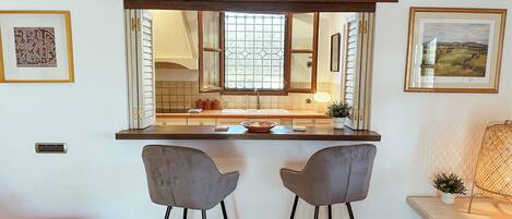 Una cocina con techo de vigas de madera, un ventanal con vistas, dos taburetes de bar y una mesita con una planta.