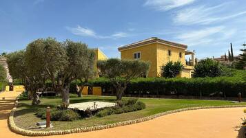 A yellow building with a tiled roof, surrounded by a well-maintained garden with various trees and shrubs.