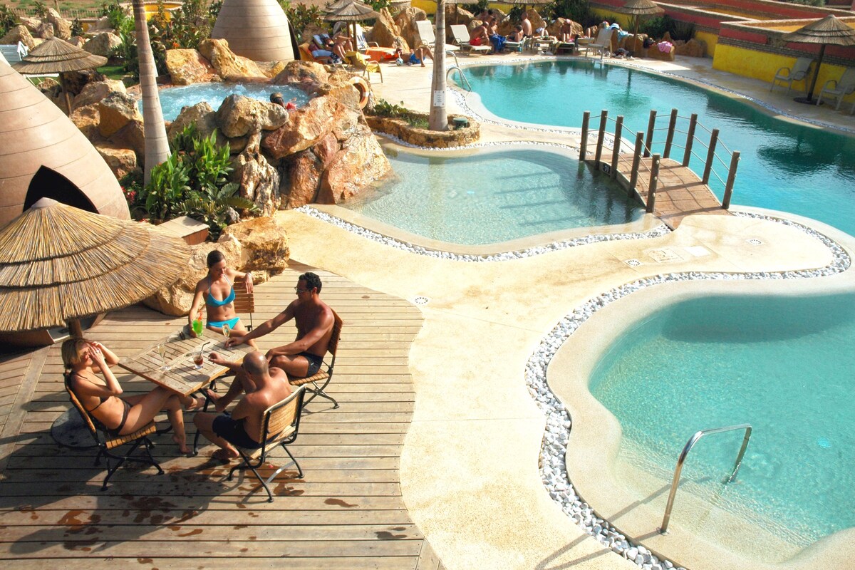 Uma área de piscina com vários elementos aquáticos, incluindo uma piscina central e piscinas menores, cercada por espreguiçadeiras e guarda-sóis.