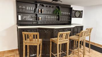 Zona de bar con barra, tres sillas de madera y estantes con vasos y una planta.