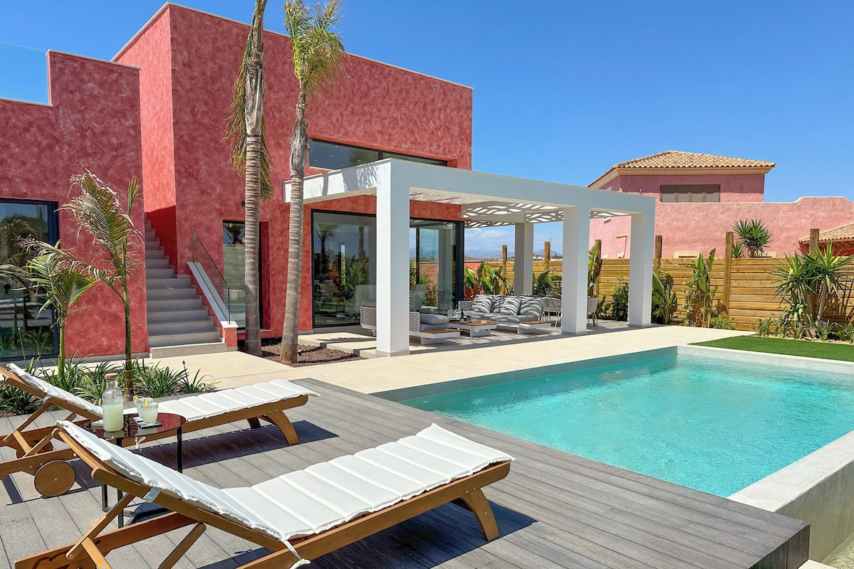 Un área moderna junto a la piscina con sillones de descanso, una pérgola y vistas a un edificio rojo.