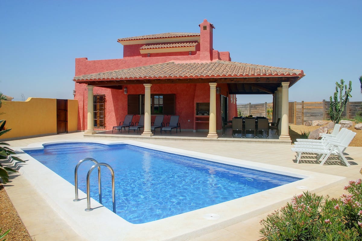Una piscina con un porche techado, un edificio rojo con techo de tejas y un paisaje desértico al fondo.