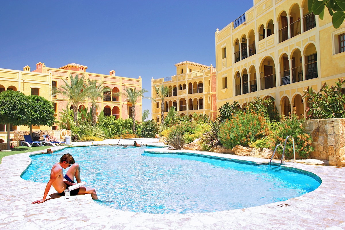 A swimming pool with a stone deck, surrounded by buildings with arched windows and balconies.