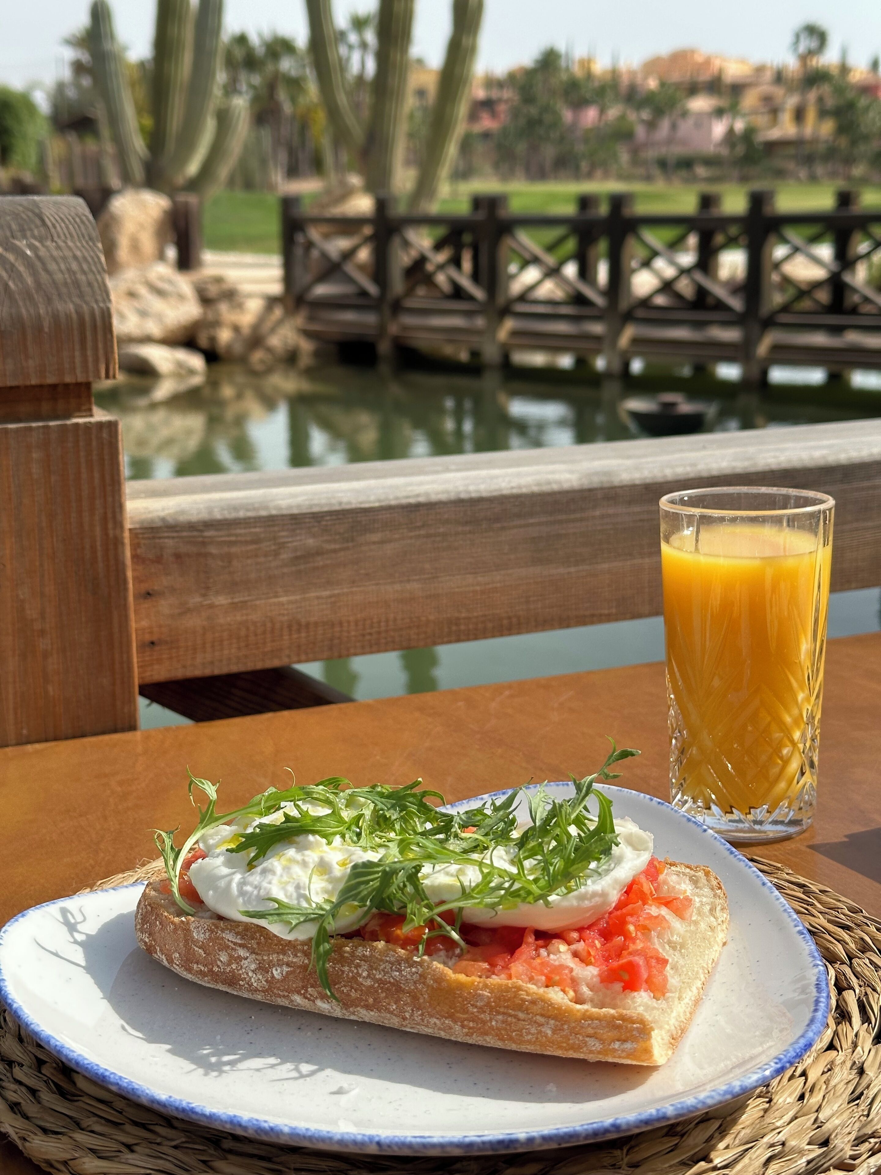 Un repas composé d’un sandwich servi sur une assiette, d’un verre de jus d’orange et d’une vue sur une étendue d’eau, avec un pont en bois en arrière-plan.