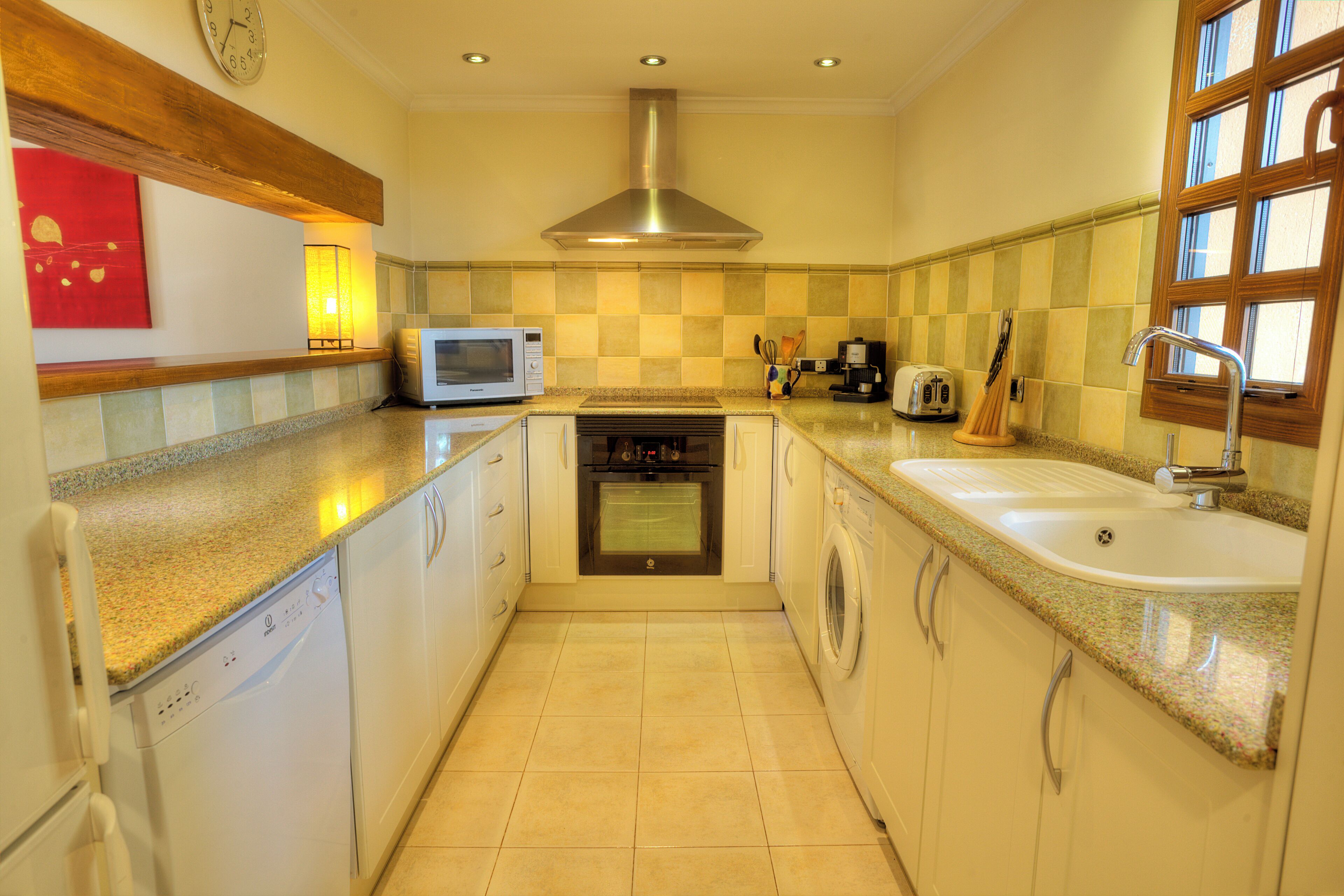A compact kitchen with a microwave, oven, washing machine, sink, and a window with blinds.