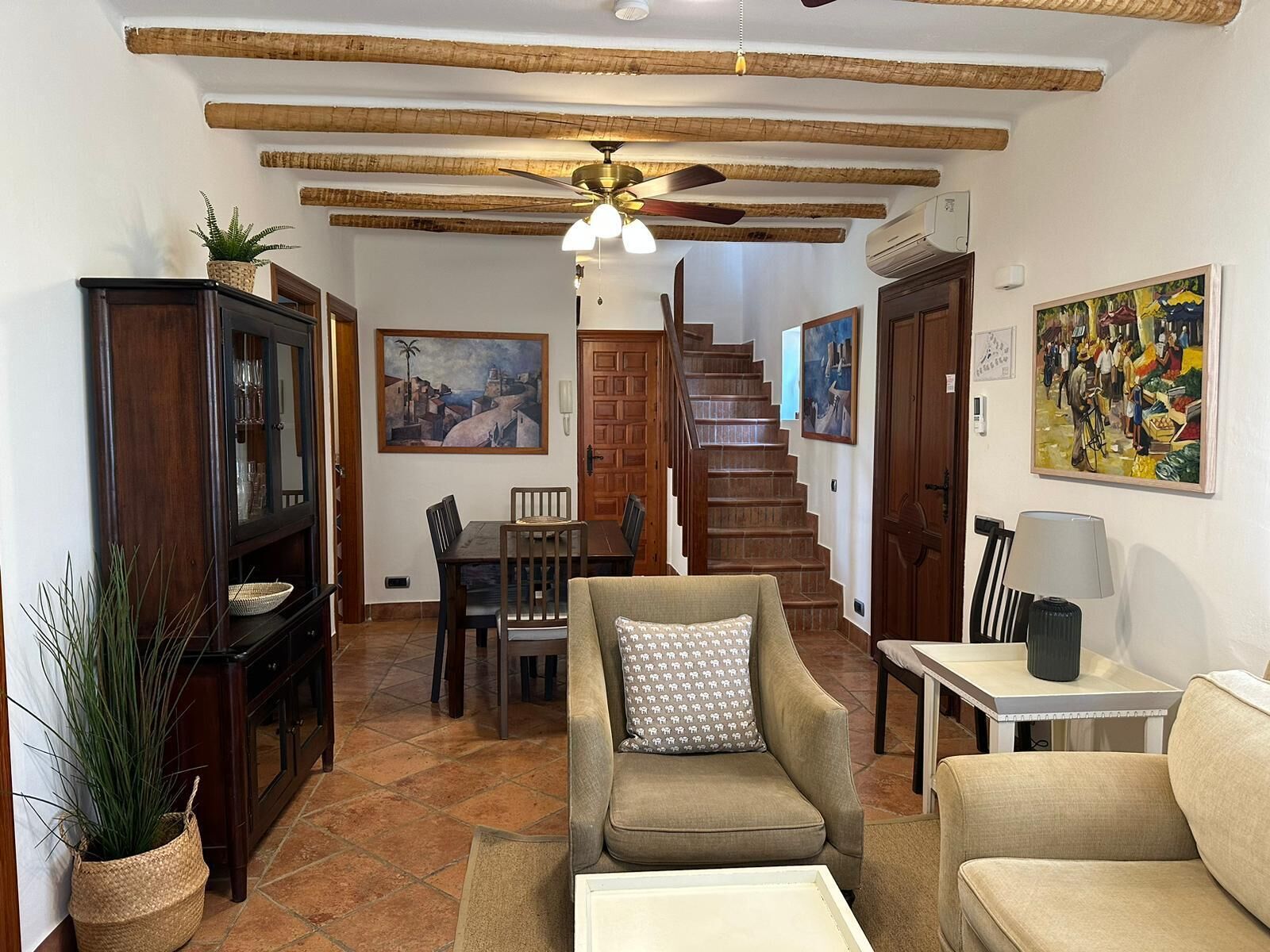 A cozy living room with a sofa, armchair, and dining table. There is a staircase leading to another level, and artwork on the walls.