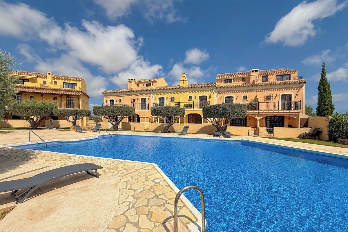 A swimming pool with a stone deck, surrounded by a large building with multiple balconies and a clear blue sky.