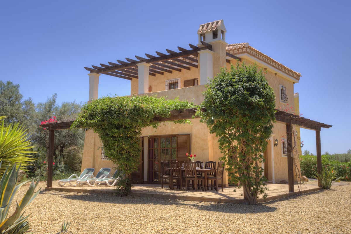 Un edificio de dos pisos con una pérgola, mobiliario exterior y un jardín.