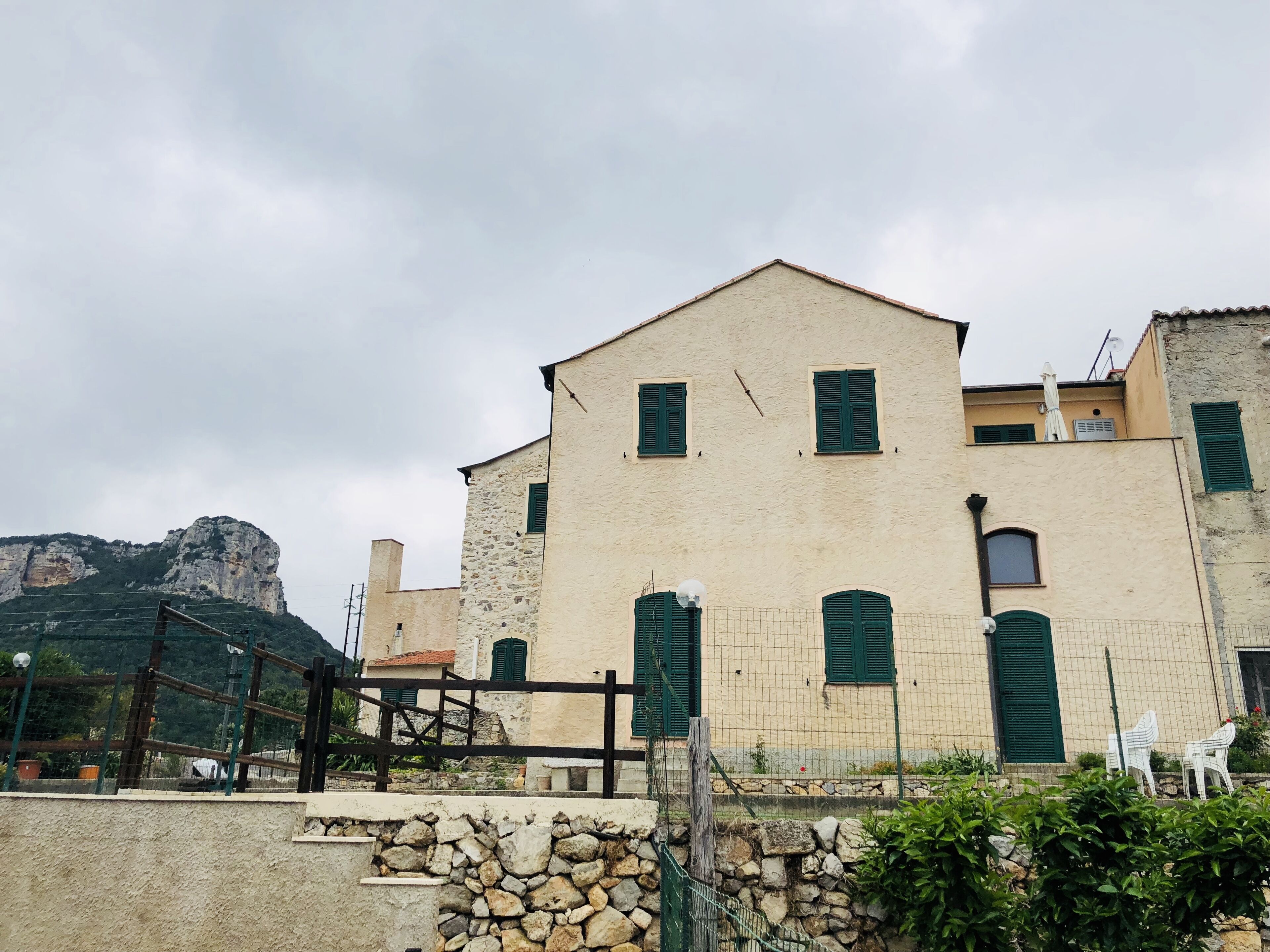 Un edificio de dos pisos con muros de piedra y contraventanas verdes, un muro de piedra en primer plano y montañas al fondo.