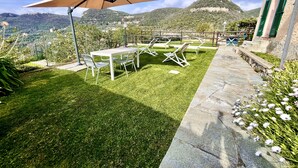 Eine Terrasse mit Tisch und Stühlen, einem großen Sonnenschirm und einem gepflasterten Weg, der zu einem Gebäude mit grünen Fensterläden führt.