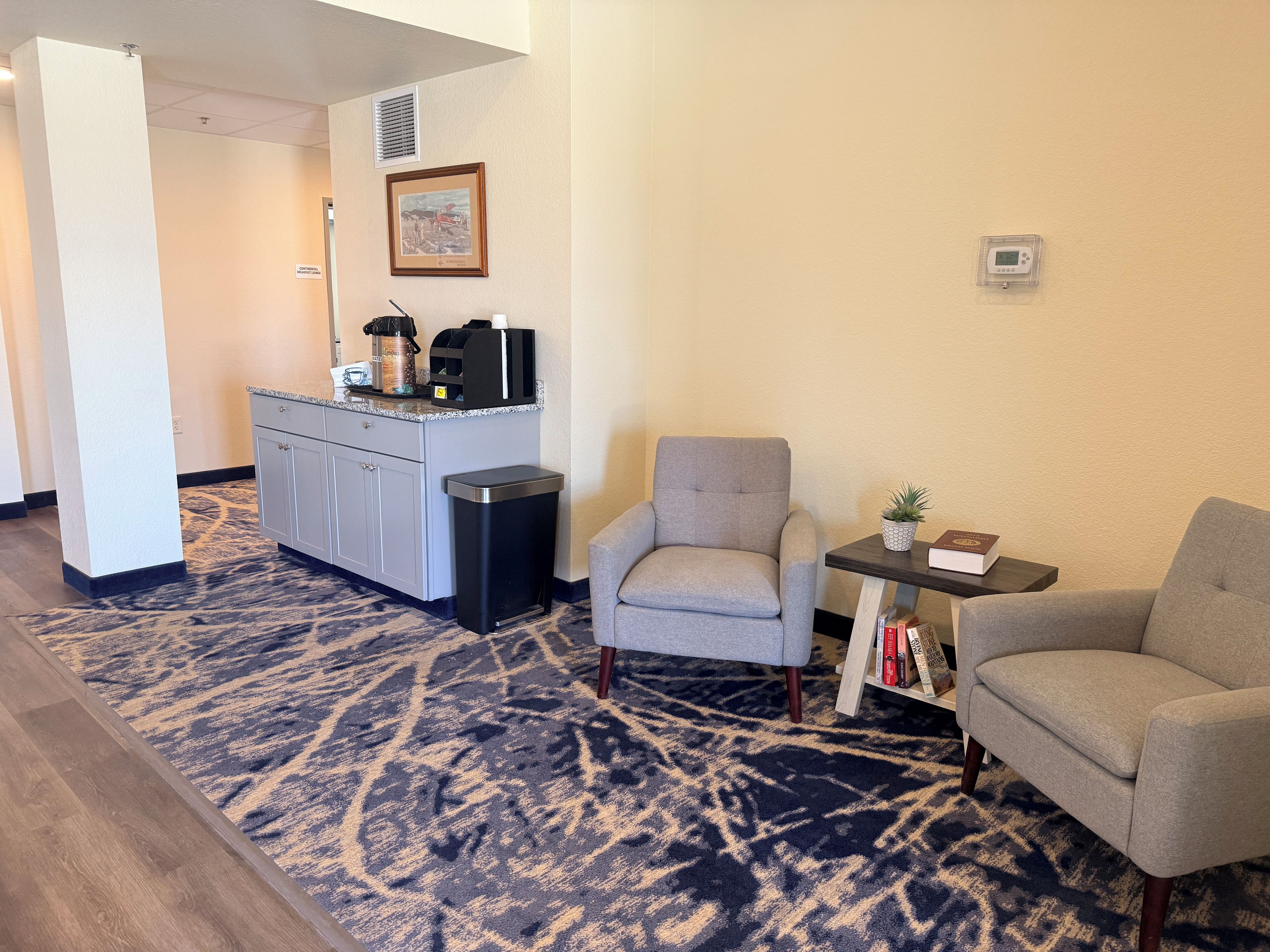 A hotel room with a kitchenette, a grey armchair, a small table with a plant and books, and a patterned carpet.