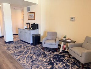 A hotel room with a kitchenette, a grey armchair, a small table with a plant and books, and a patterned carpet. - Iron Creek Inn & Suites (Upton)