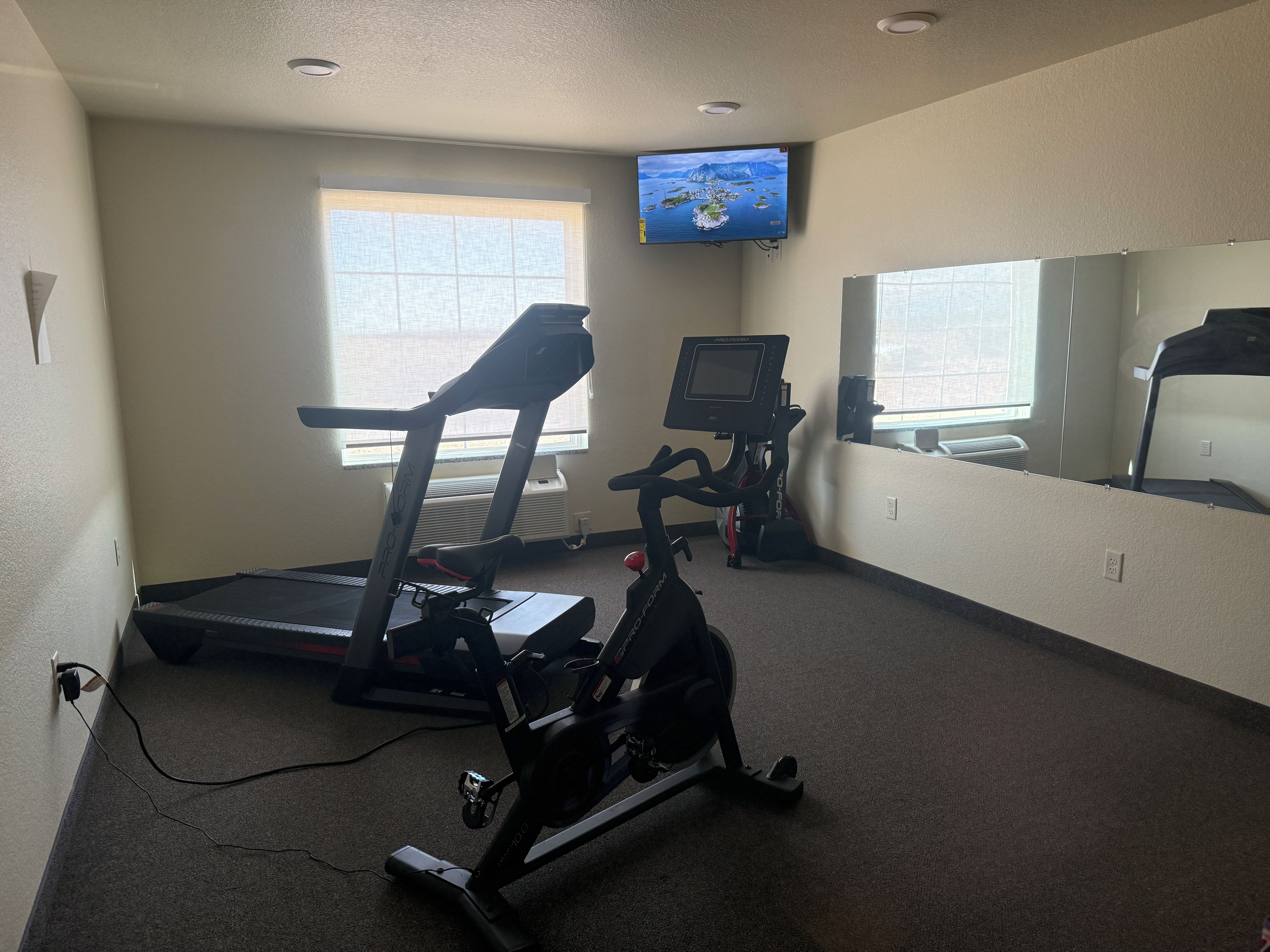 Un gimnasio con equipamiento de ejercicio, un televisor montado en la pared y un ventanal que proporciona luz natural.