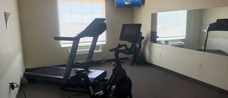 Un gimnasio con equipamiento de ejercicio, un televisor montado en la pared y un ventanal que proporciona luz natural.