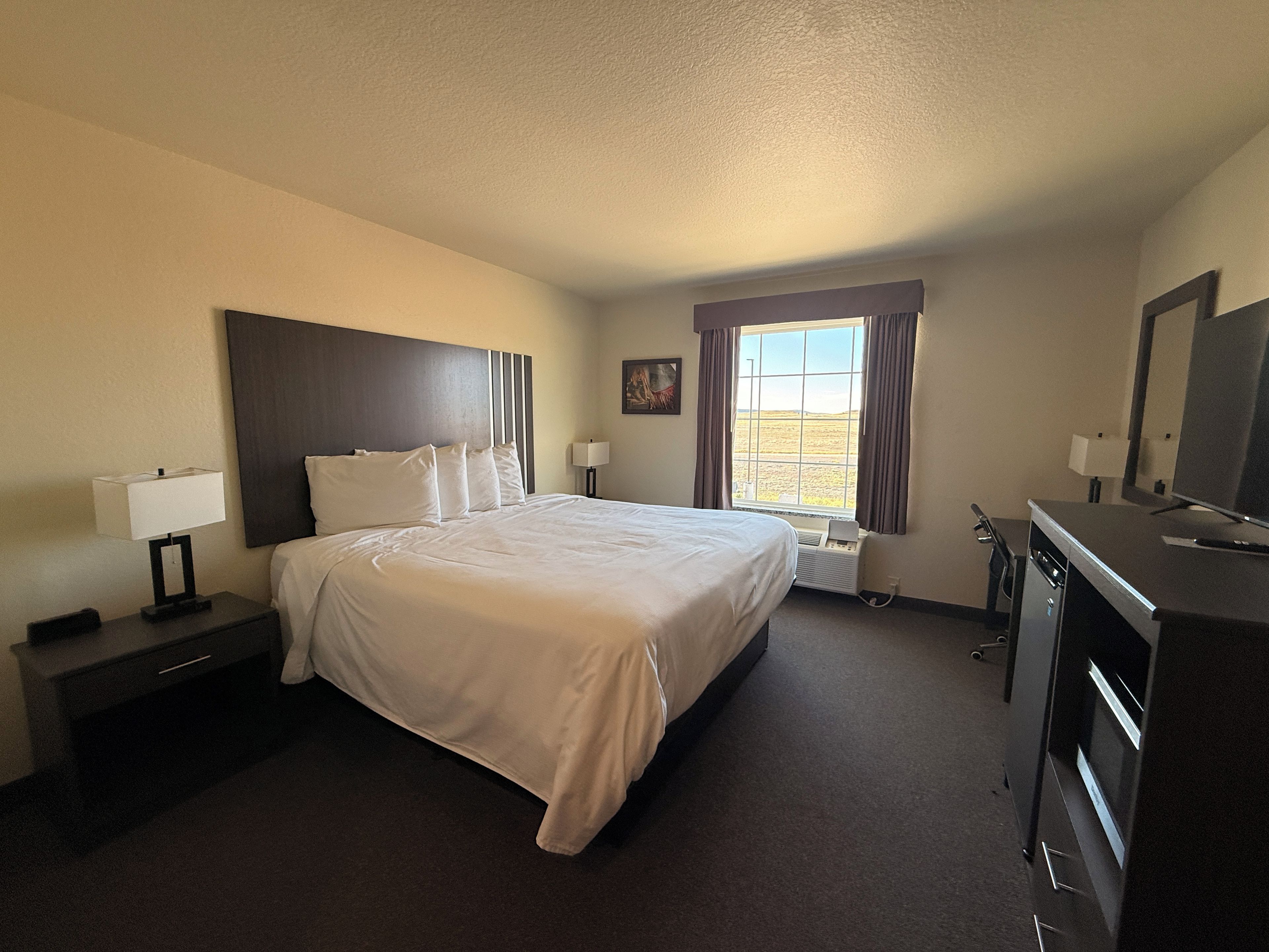 A hotel room with a large bed, a desk with a computer, a dresser, and a window with curtains.