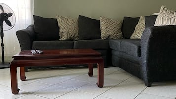 A living room with a sectional sofa, a wooden coffee table, a standing fan, and an air conditioning unit on the wall.