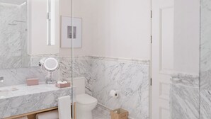 Um banheiro moderno com parede de mármore, box de vidro, vaso sanitário branco e toucador com espelho e dispensador de sabonete.