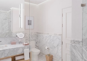 Un baño moderno con pared de mármol, mampara de ducha de vidrio, inodoro blanco y tocador con espejo y dispensador de jabón.