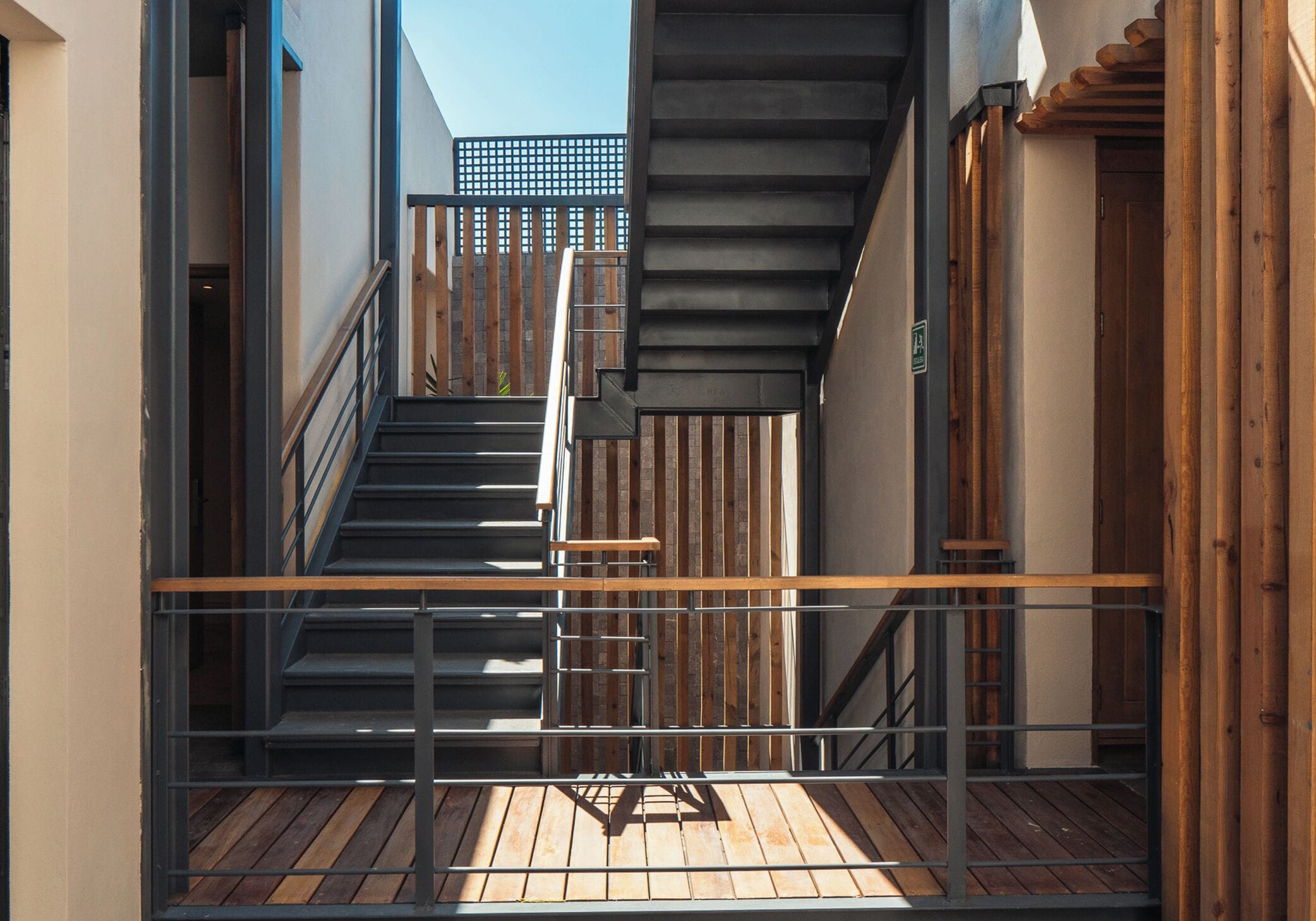 A narrow outdoor corridor with a wooden deck, metal railing, and a staircase leading to an upper level.