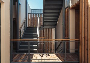 Un étroit couloir extérieur doté d’une terrasse en bois, d’une rambarde en métal et d’un escalier menant à un niveau supérieur.