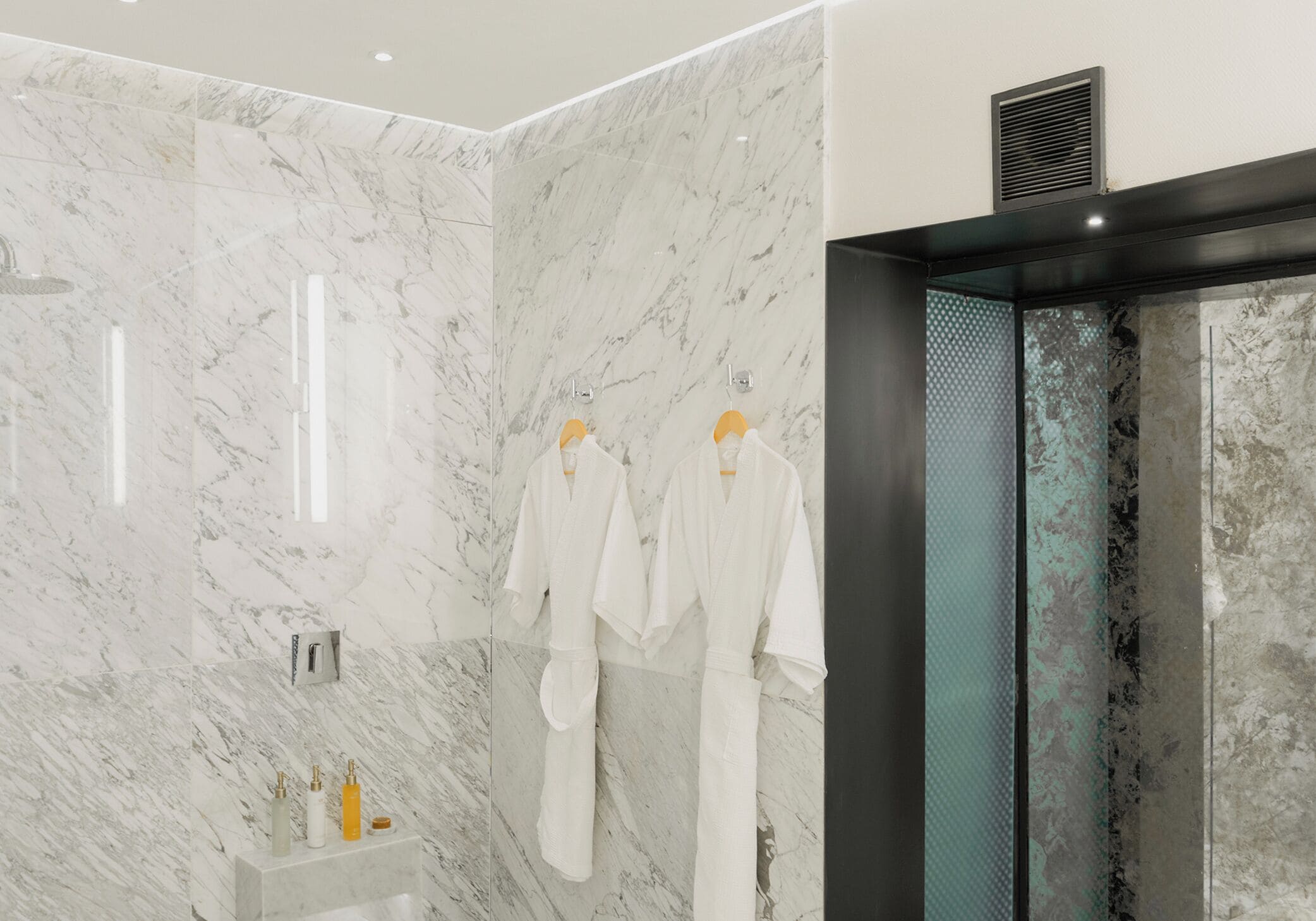 A modern bathroom with marble walls, a glass shower enclosure, and two white robes hanging on hooks.