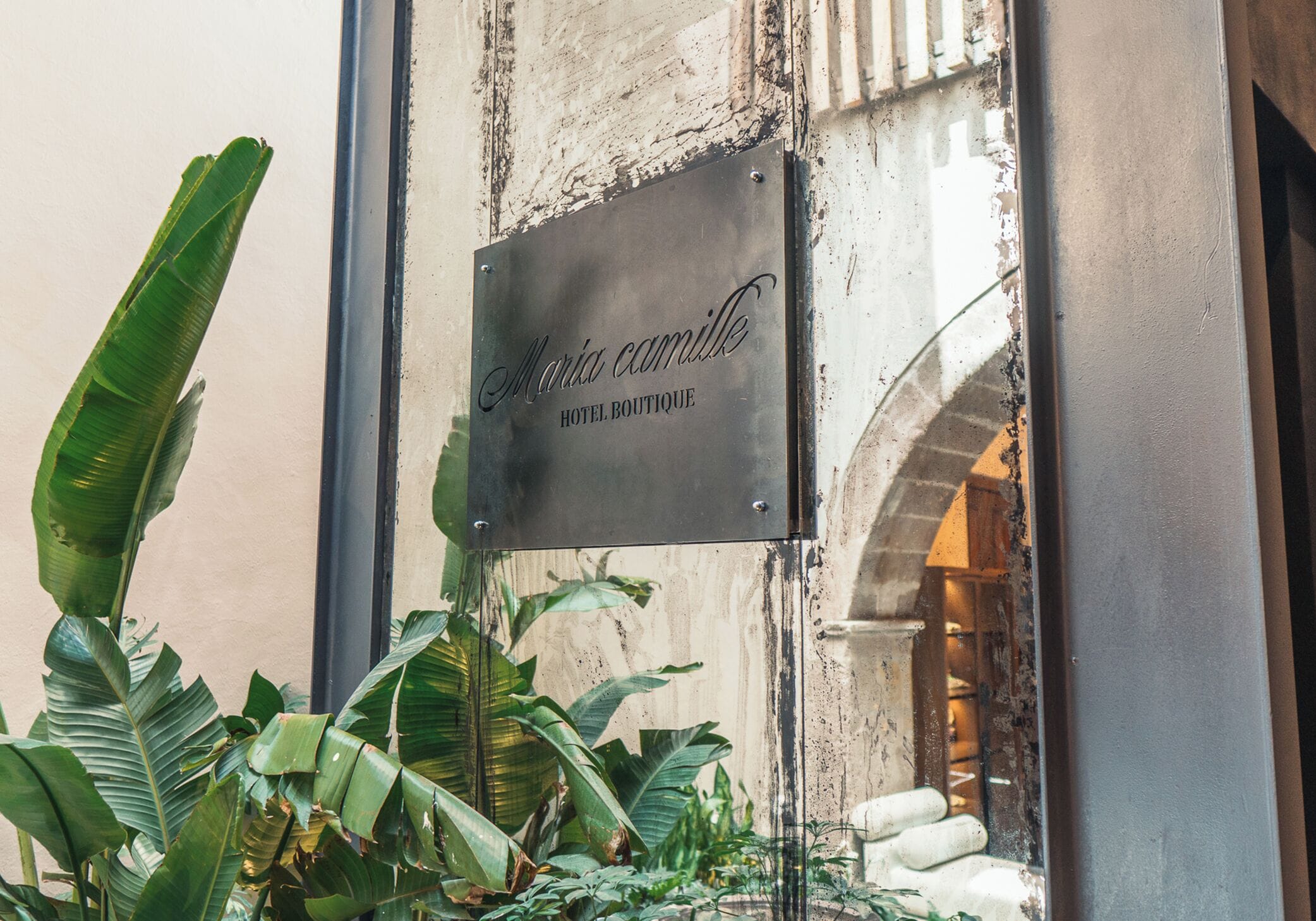 Un letrero metálico con la inscripción 'Maria Camilla HOTEL BOUTIQUE' grabado, sujeto a un edificio de fachada desgastada y un arco visible.