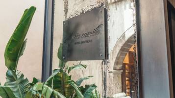 Ein Metallschild mit der eingravierten Aufschrift 'Maria Camilla HOTEL BOUTIQUE', angebracht an einem Gebäude mit verwitterter Fassade und einem sichtbaren Bogen.