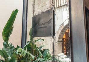 Ein Metallschild mit der eingravierten Aufschrift 'Maria Camilla HOTEL BOUTIQUE', angebracht an einem Gebäude mit verwitterter Fassade und einem sichtbaren Bogen.