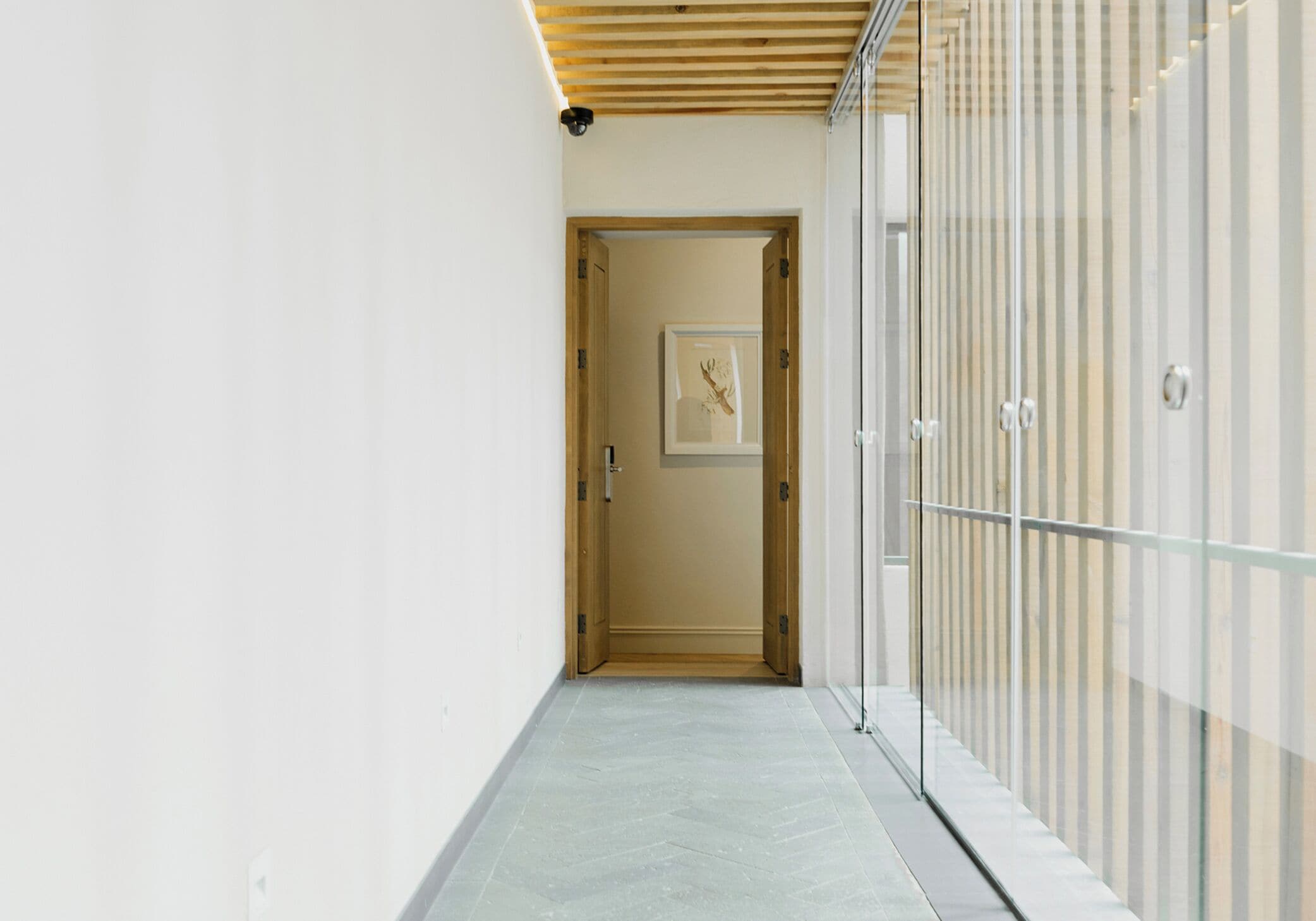 Un couloir avec un plafond en bois, une porte ornée d’un tableau encadré et une cloison vitrée sur la droite.