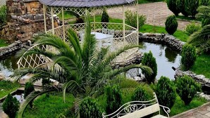 A gazebo with a tiled roof, surrounded by a pond, palm trees, and a bench. - Hotel Oasis Villa (Tsqaltubo)