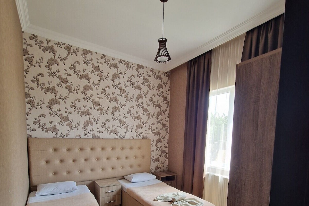 Ein Hotelzimmer mit einem großen Bett, einem kleineren Bett, Holzboden und einer Deckenleuchte.