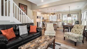 A spacious living area with a black leather sofa, a patterned coffee table, and a white armchair. A kitchen with white cabinets and a dining area with a wooden table and chairs are visible. - Family-Friendly 3BR Condo Grand Haven 27 (Grand Haven)