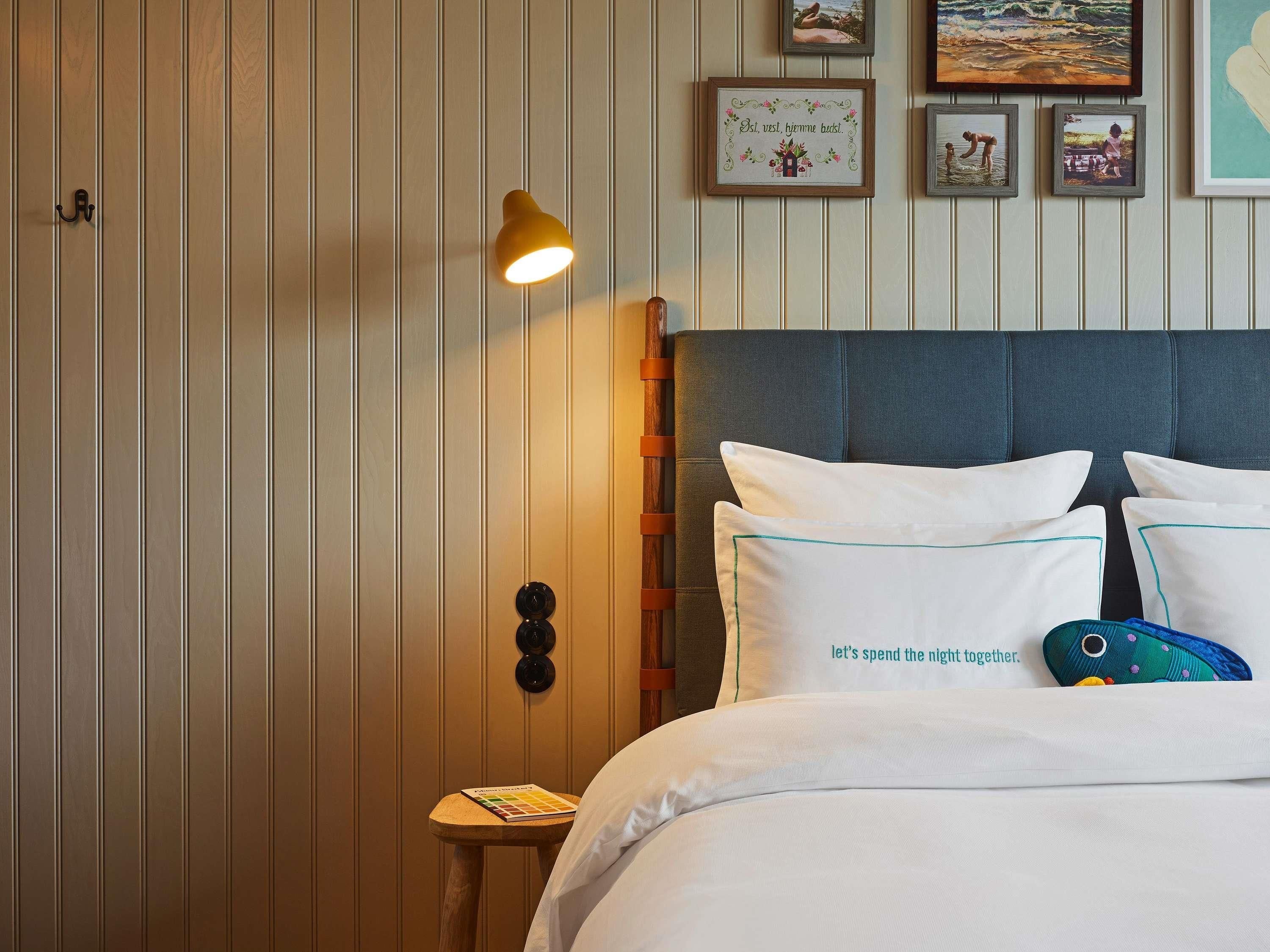 A hotel room with a bed, a bedside table with a book, a wall-mounted lamp, and framed pictures on the wall.