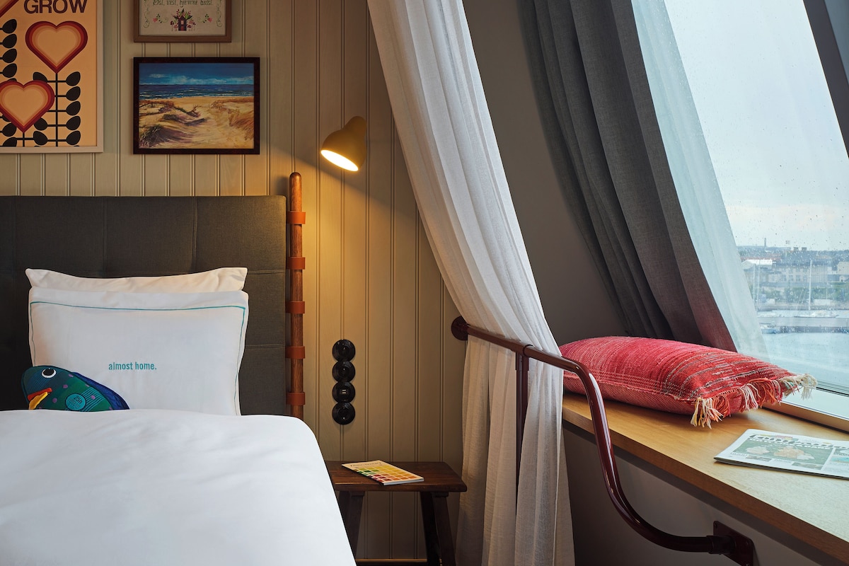 Ein Hotelzimmer mit Bett, einem kleinen Tisch mit einem Buch, einem Fenster mit Vorhängen und einer Wand mit gerahmten Bildern.