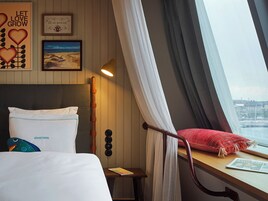 A hotel room with a bed, a small table with a book, a window with curtains, and a wall with framed pictures.