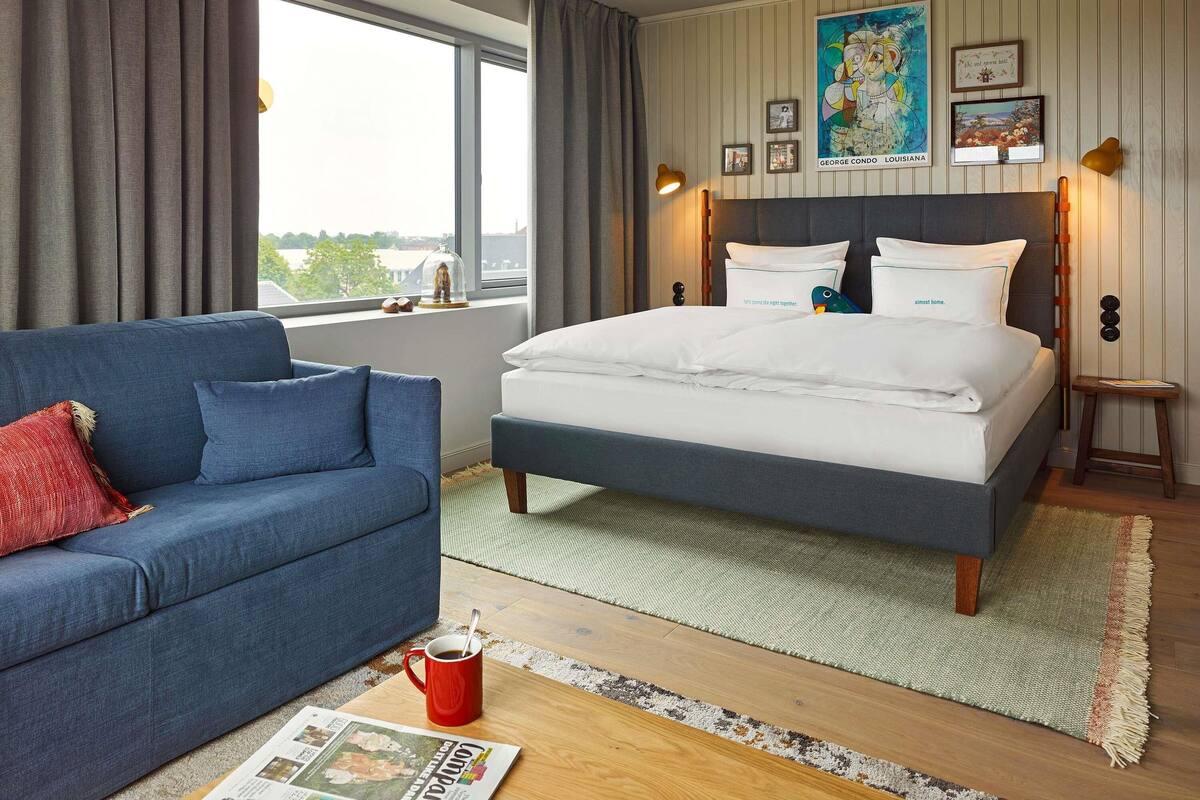 A bedroom with a bed, a blue sofa, a window with curtains, and a view of the city.