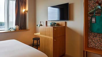 A hotel room with a bed, a wooden desk with a telephone, a television mounted on the wall, a window with curtains, and a wall with a patterned wallpaper.