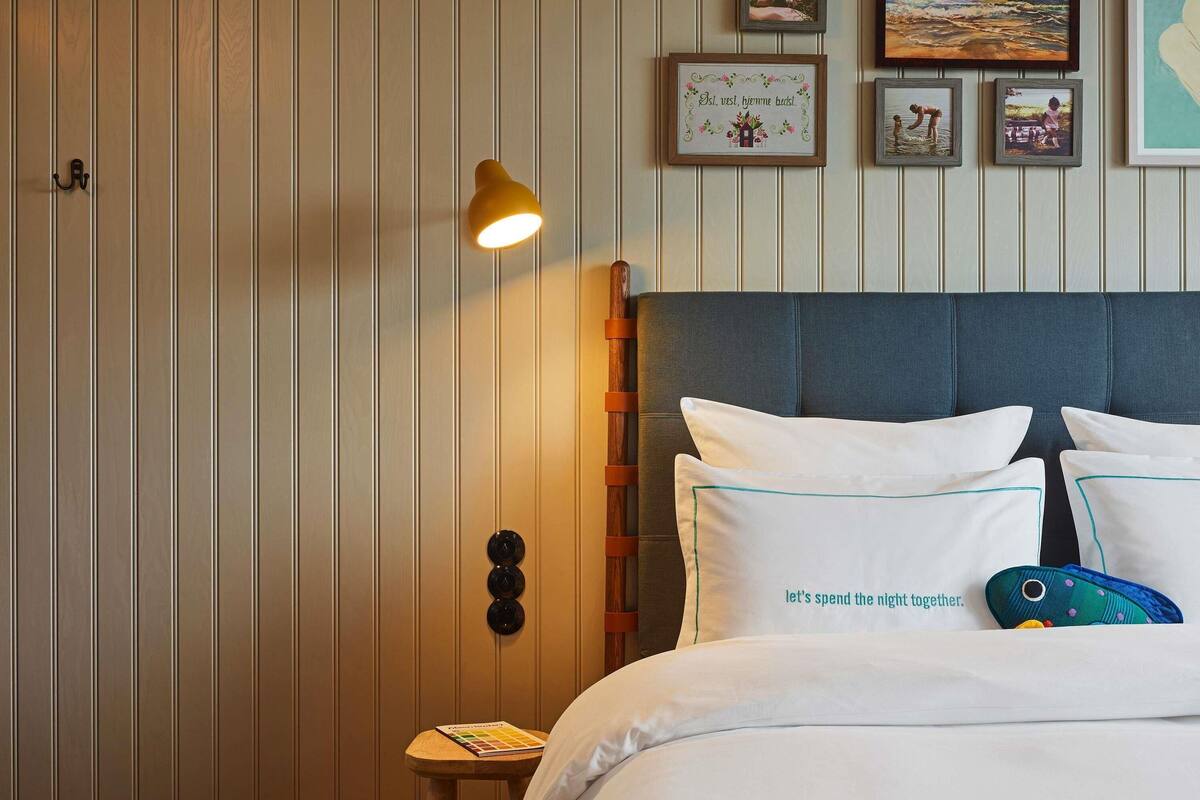 A hotel room with a bed, a bedside table with a book, a wall-mounted lamp, and framed pictures on the wall.