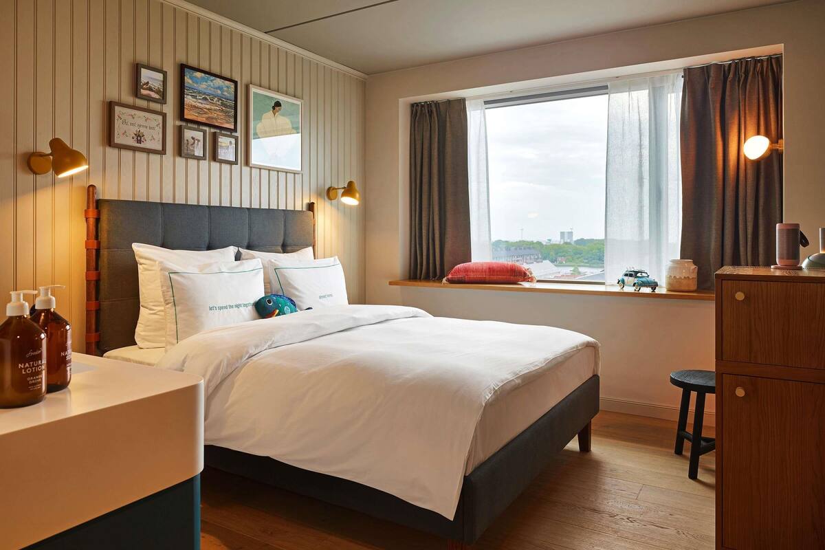 A neatly made bed with white linens, a wooden headboard, and a view of a cityscape through a window with curtains.