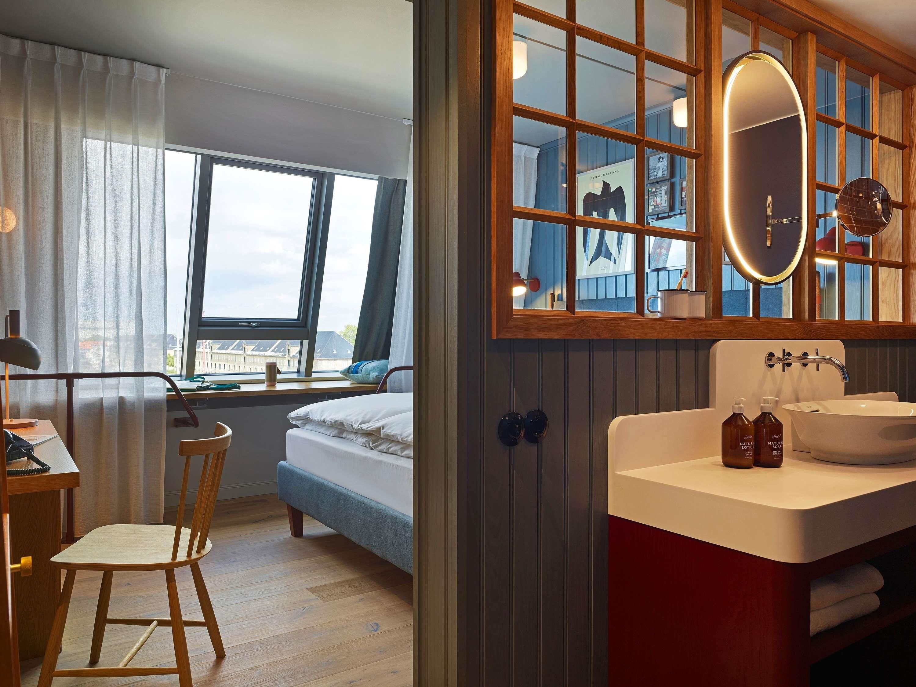 Habitación de hotel con cama, escritorio y silla, ventana con cortinas, baño con lavamanos y espejo, y vistas a la ciudad.